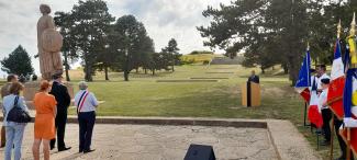 Porte-drapeaux, public et un élu derrière un pupitre dans un cadre verdoyant, un monument en arrière-plan.